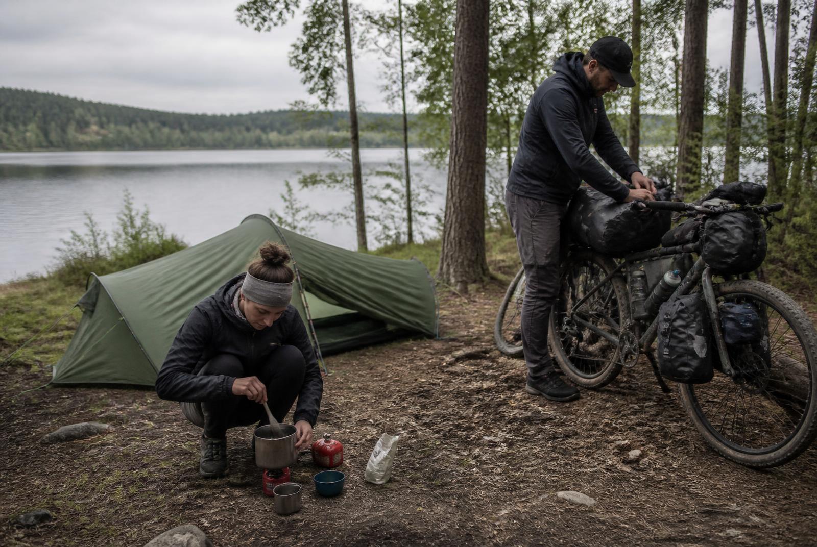 Zwei Radreisende bereiten an einem See ihr Lager und Essen vor
